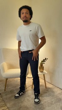 Bela majica z okrolim izrezom, temno modre hlače in črno-bele superge. Teksturirana svetla ozadje z belim žametnim stolom in vazo.