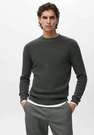 Joven con cabello oscuro que lleva un suéter de canalé gris oscuro sobre una camisa blanca, combinado con pantalones grises, manos en los bolsillos, fondo neutro.