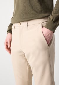 Beige trousers with a smooth texture, flat front, and side pockets. Simple design featuring a button closure. Worn with a dark green sweater.