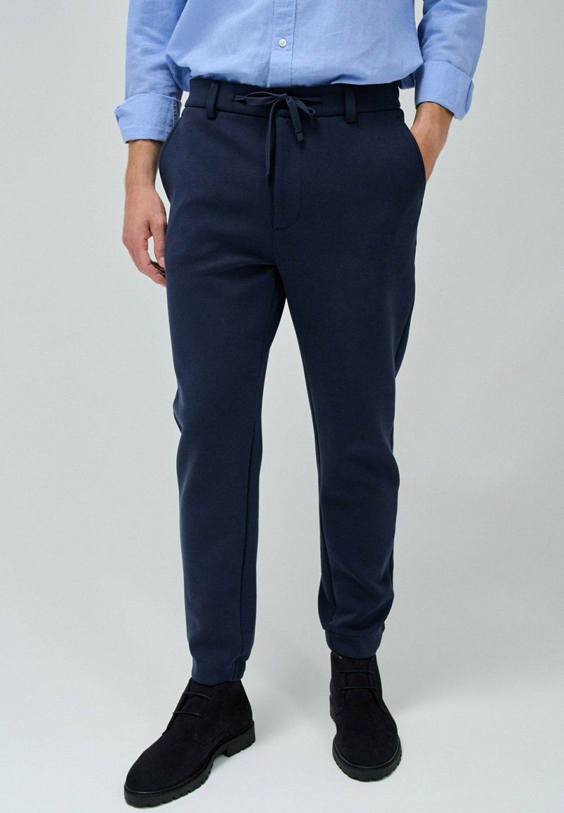 Pantalones de chándal azul marino hechos de una tela suave, con cintura ajustable, pierna estrecha y bolsillos laterales. Combinados con botas negras.