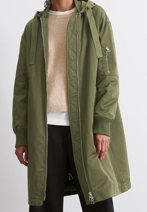 Person wearing long olive green hooded jacket with front zipper, beige sweater, white shirt, and black pants against plain background.