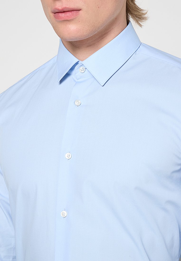 Uomo che indossa una camicia elegante azzurro chiaro con colletto a punta, mostrando solo la parte inferiore del viso e la parte superiore del torso.