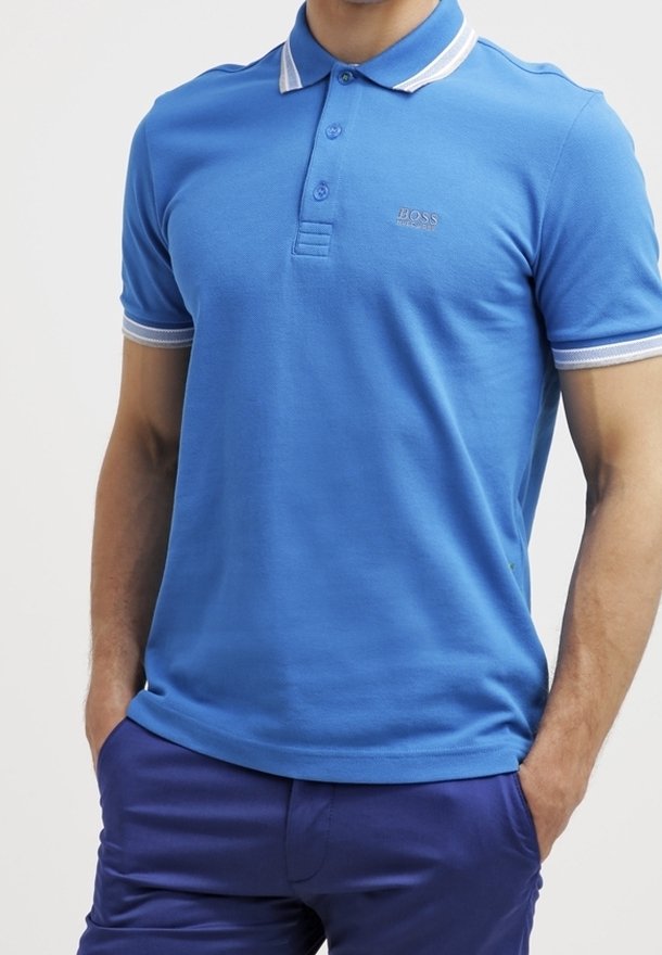 Homme portant un polo bleu ajusté avec une bordure blanche sur le col et les manches, associé à un pantalon bleu foncé, les mains dans les poches.