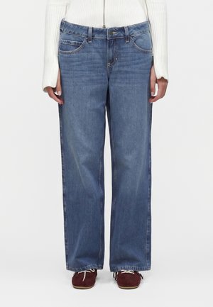 Person, die gerade geschnittene blaue Jeans, einen weißen Rippstrickpullover mit Reißverschluss und dunkelrote Schnürschuhe trägt, steht vor einem schlichten Hintergrund.