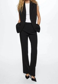 Gilet noir ajusté avec un seul bouton et des détails de poches, associé à un pantalon droit assorti, porté par-dessus un haut blanc et des talons noirs.
