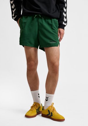 Groene sportshorts met een trekkoord in de taille, voorzakken en branding. Gecombineerd met gele sportschoenen en witte sokken met zwarte strepen.