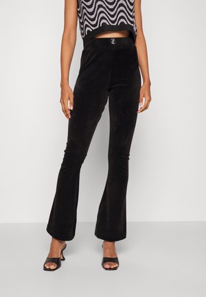 Vrouw draagt zwarte uitlopende broek, een zwart-wit top met golvend patroon en open-teen zwarte sandalen met hak, staand tegen een effen achtergrond.