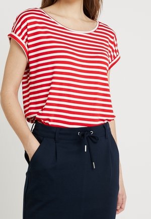 Vrouw draagt een rood-wit gestreept T-shirt met korte mouwen, ingestopt in een zwarte rok met trekkoord in de taille, staat met één hand in de zak.