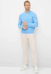 Lyseblå langærmet sweater med rund halsudskæring, kombineret med lyse beige chinos og hvide sneakers, stående mod en ensfarvet baggrund.