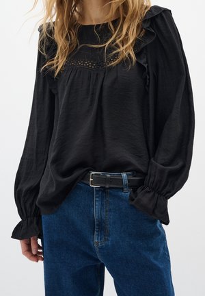 Vrouw draagt een zwarte blouse met lange mouwen met ruches en een blauwe spijkerbroek, vastgemaakt met een zwarte riem tegen een effen achtergrond.