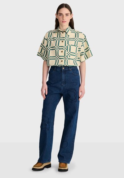 Femme portant une chemise beige à manches courtes avec un motif à carreaux verts, un jean taille haute bleu foncé avec des patchs et des bottes marron clair, debout sur fond blanc.