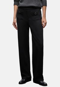 Pantaloni neri a gamba larga realizzati in tessuto liscio, con due tasche frontali decorate da fibbie e una cintura elastica.