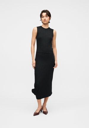 Žena stojaca v čiernom tielku bez rukávov a zodpovedajúcej dlhej sukni, obutá v hnedých lodičkách so špicatou špičkou, na jednoduchom svetlom pozadí.