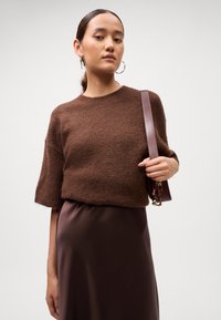 Žena v hnedom textúrovanom krátkom rukávovom svetri a tmavohnedej saténovej sukni, ktorá drží zladenú hnedú kabelku na pleci.