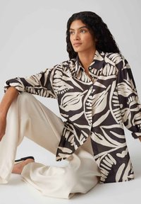 Camicia morbida realizzata in tessuto leggero con una base marrone scuro e un motivo floreale crema; presenta un colletto e chiusura a pulsante.
