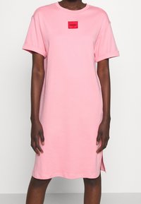 Różowa bawełniana sukienka z krótkimi, podwiniętymi rękawami i długością sięgającą powyżej kolan. Posiada małą, czerwoną łatkę z logo na klatce piersiowej.