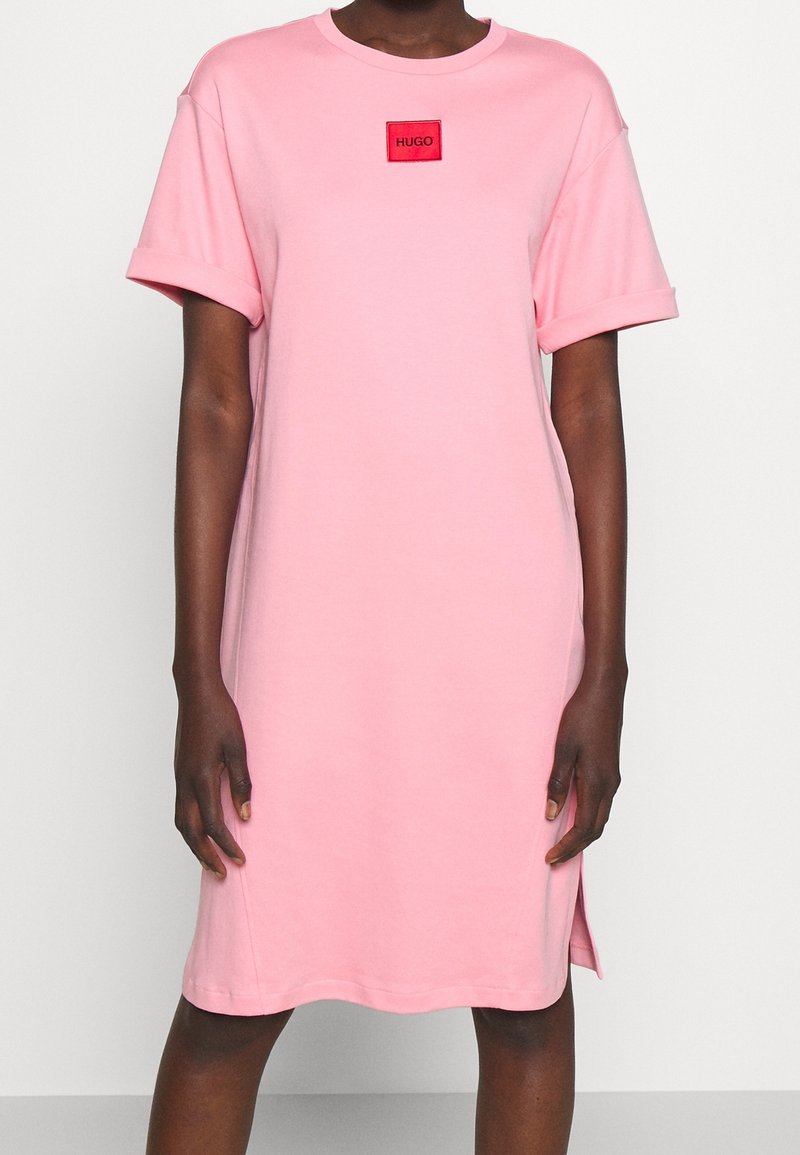 Różowa bawełniana sukienka z krótkimi, podwiniętymi rękawami i długością sięgającą powyżej kolan. Posiada małą, czerwoną łatkę z logo na klatce piersiowej.