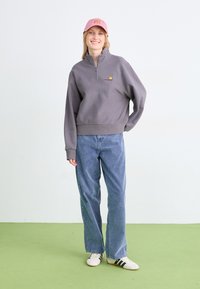 Sudadera gris con cremallera, puños de canalé, detalle del logo, combinada con jeans rectos de color azul claro y zapatillas blancas con rayas negras.