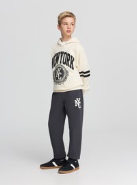 Felpa con cappuccio beige con stampa nera "NEW YORK" e strisce, abbinata a pantaloni della tuta grigi con logo bianco, completata da scarpe nere.