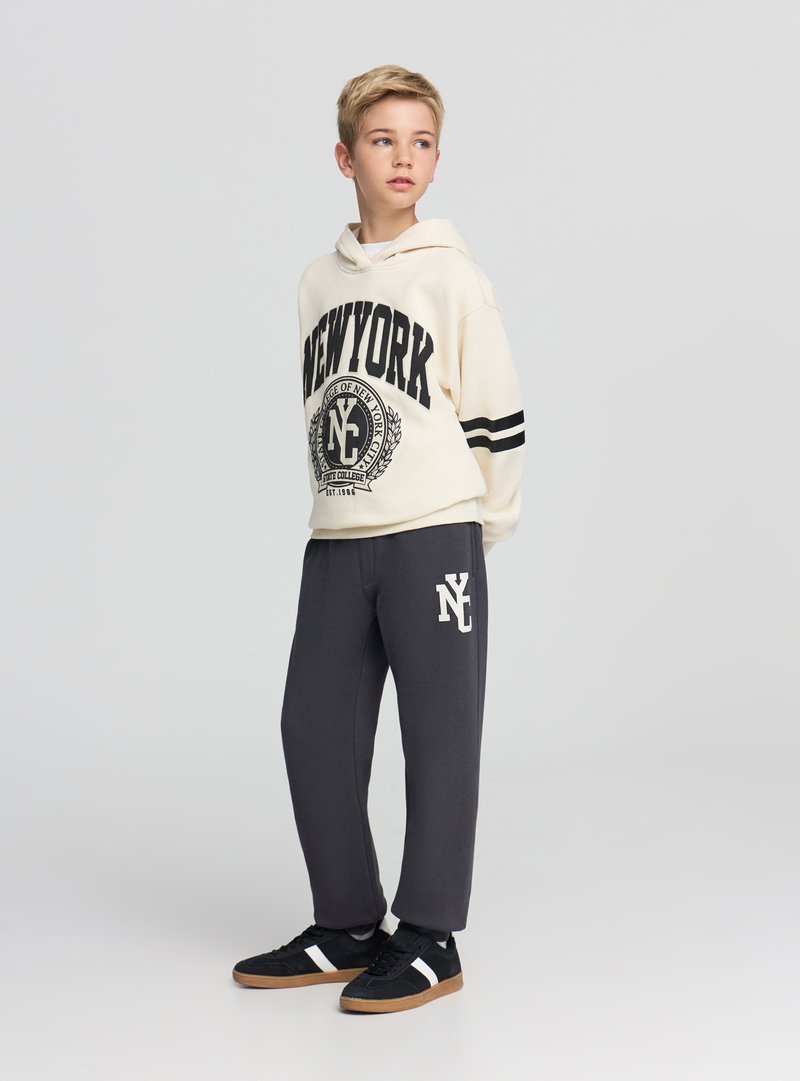 Felpa con cappuccio beige con stampa nera "NEW YORK" e strisce, abbinata a pantaloni della tuta grigi con logo bianco, completata da scarpe nere.