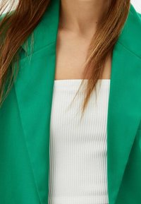 Veste verte à revers pointus portée sur un haut blanc côtelé. Le tissu de la veste semble lisse, avec un design à ouverture frontale.