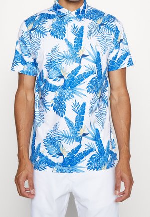 Mężczyzna w białej koszuli z krótkim rękawem z niebieskim tropikalnym wzorem liści i kwiatów, ubrany w białe spodnie, stoi na jednolitym tle.