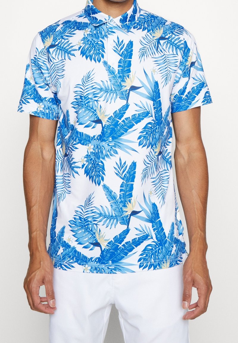 Homme portant une chemise blanche à manches courtes avec un imprimé de feuilles tropicales bleues et de fleurs, associée à un pantalon blanc, debout devant un fond uni.
