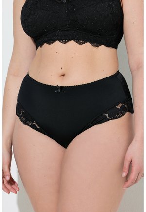 Woman wearing black high-waisted underwear with lace details and a matching black lace bralette, standing against a plain background.