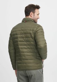 Olivgrüne Pufferjacke mit horizontalen Nähten, hohem Kragen und Seitentaschen. Hat einen rot-weißen Akzent auf dem Rücken.