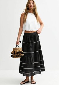 Jupe noire à volants avec des accents blancs, associée à un top court blanc. Les accessoires incluent un sac en osier avec des bords volantés et des tongs.