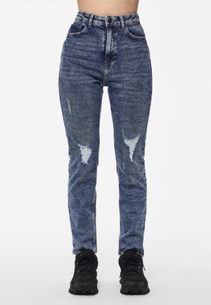 Kvinde iført højtaljede slidte blå jeans og sorte sneakers, stående mod en ensfarvet hvid baggrund.