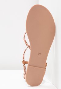 Semelle d'une sandale à brides tressées rose et beige, taille 37 indiquée, posée en angle contre une surface d'angle blanche.