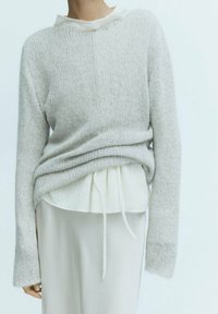 Personne portant un pull en tricot gris clair par-dessus un chemisier blanc à manches longues avec une taille nouée, associé à une jupe de couleur claire.