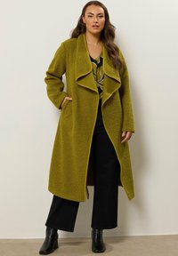 Abrigo largo color verde oliva con una tela texturizada y suave. Presenta un collar amplio y abierto, así como bolsillos. Se lleva sobre una blusa estampada y pantalones negros.