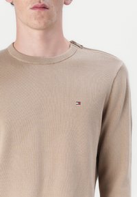 Beige hosszú ujjú pulóver bordás kötött anyagból. Kerek nyakkivágással rendelkezik, és a mellkason egy kis logó címke található piros, fehér és kék színekkel.