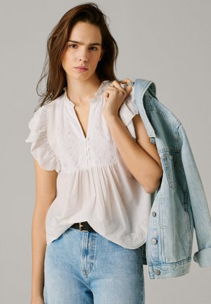 Młoda kobieta w białej bluzce z ażurowym wzorem i jasnoniebieskich dżinsach trzyma na ramieniu kurtkę dżinsową na jednolitym tle.