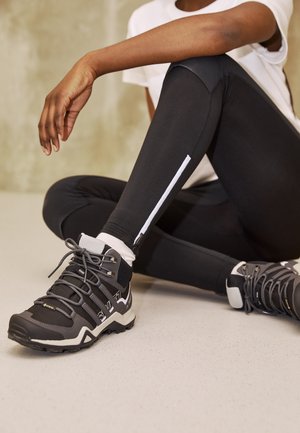 Persoon zit met gekruiste benen en draagt zwarte leggings met een witte rits en zwarte hoge wandelschoenen met grijze veters.