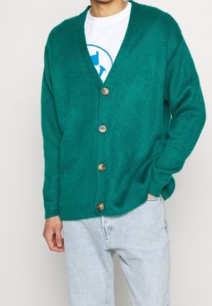 Homme portant un cardigan boutonné bleu sarcelle sur un t-shirt blanc graphique avec un jean bleu clair, main dans la poche, debout devant un fond uni.