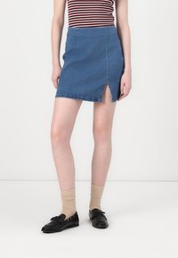Denim mini szoknya egyenes szabással, elülső hasítékkal, kék színben, finom varrási részletekkel, fekete cipőkkel és zoknikkal párosítva.