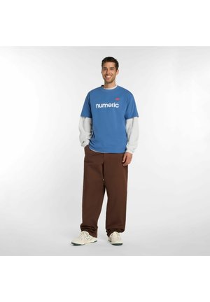 Lächelnder Mann mit blauem „numeric“-T-Shirt über weißen Langarmshirts, brauner Hose und weißen Turnschuhen, stehend vor weißem Hintergrund.