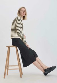 Gestreifter Creme- und schwarzer Pullover, lockere schwarze Culottes und klobige schwarze Schuhe. Modell sitzt auf einem Holzstuhl vor einem hellen Hintergrund.