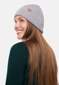 Gray knitted beanie with ribbed texture and a small brown leather label. Worn slightly back on the head, featuring long brown hair in the background.