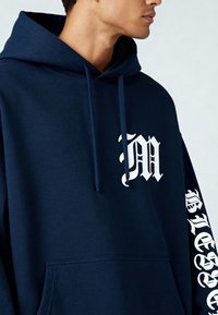 Navyblauwe katoenen hoodie met een voorvak, voorzien van een witte letter "M" op de borst en witte tekst langs de mouw. Zachte textuur.