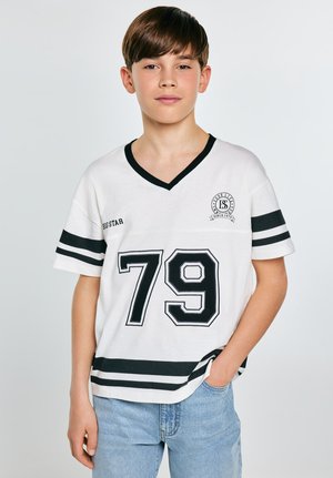 Chłopiec o ciemnych włosach ubrany w biały sportowy t-shirt z czarnymi paskami i numerem 79, pozujący z jedną ręką w kieszeni na neutralnym tle.