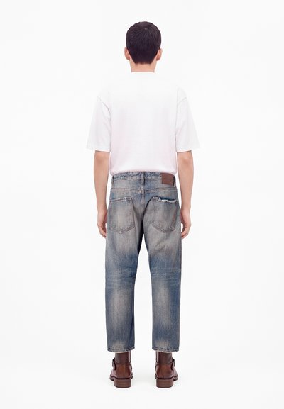 Homme portant une chemise blanche à manches courtes, un jean bleu délavé et des bottes en cuir marron, debout dos à un fond blanc.