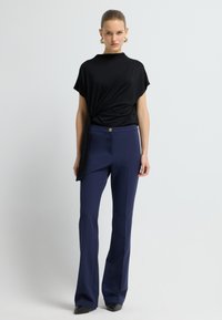 Svart draperad topp med korta ärmar, matchad med blå utsvängda byxor med knäppning. Slätt tyg, minimal detaljering.