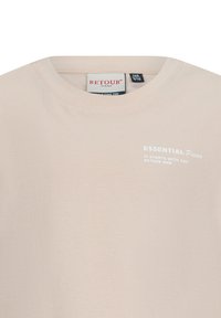 Beżowa koszulka z miękkiego materiału, z okrągłym dekoltem i małym białym nadrukiem na froncie, na którym widnieje napis "ESSENTIAL Pieces".