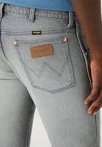 Jeans de mezclilla gris con un parche de cuero con el logo de Wrangler en el bolsillo trasero, remaches de cobre y un diseño ajustado.