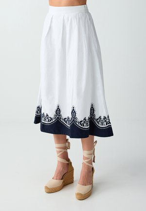 Femme portant une jupe midi blanche à ourlet brodé bleu marine et des sandales compensées espadrilles beiges à liens autour de la cheville, debout devant un fond uni.