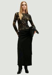 Schwarzes Langarmshirt mit einem strukturierten, mehrfarbigen Muster, kombiniert mit einem schwarzen Wickelrock. Das Model trägt kniehohe schwarze Stiefel und hält eine Tasche.